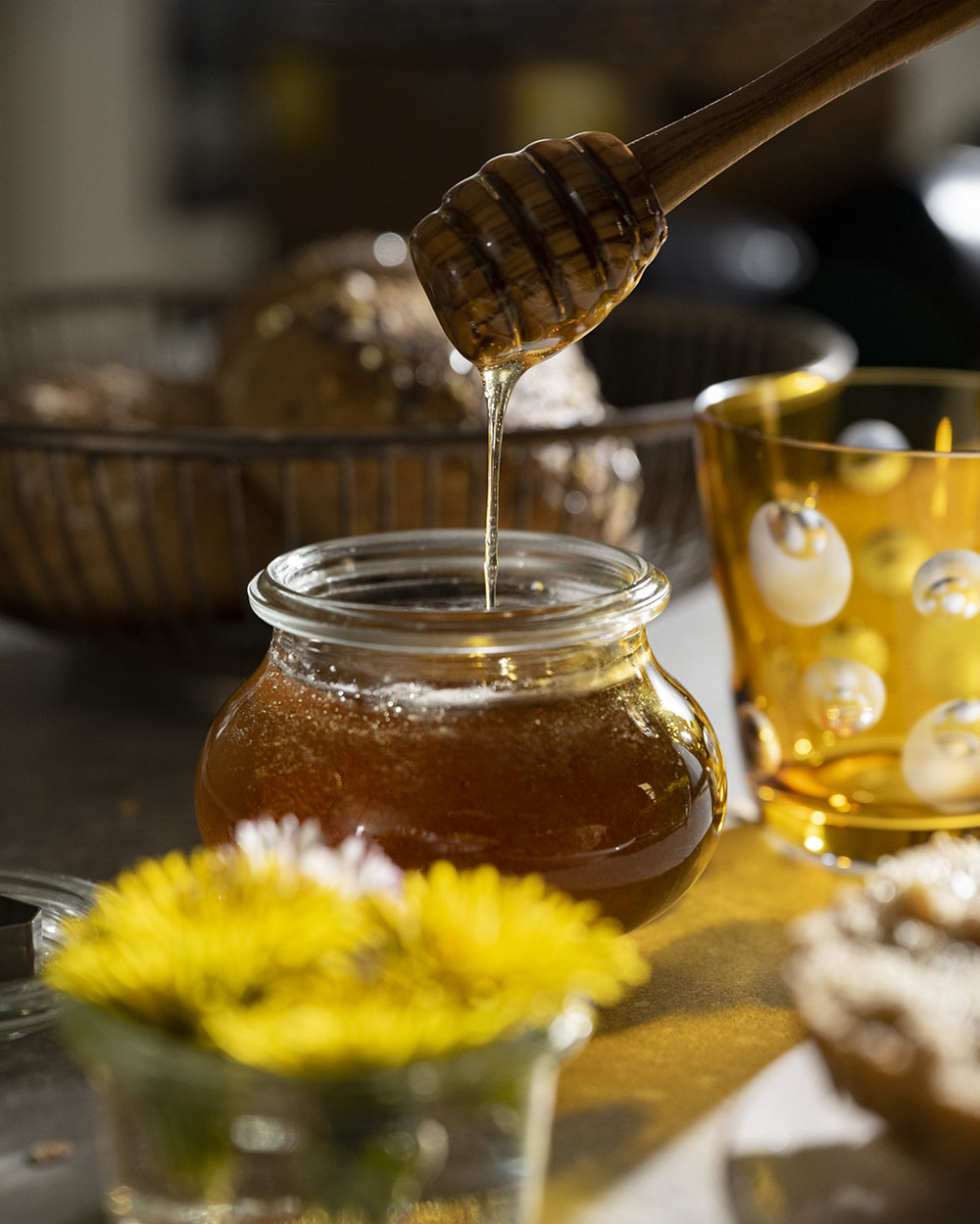 Wir tauchen einen Honiglöffel in ein Glas mit unserem selbst gemachten Löwenzahnhonig. Das Glas steht auf einem Frühstückstisch. Im Hintergrund ist ein Korb mit Brötchen zu sehen.
